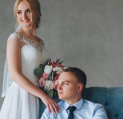 A lovely bride is standing and touching a stylish groom on the shoulder, he is sitting on a blue sofa in a grey studio. Wedding portrait of beautiful newlyweds. Wedding interior. 