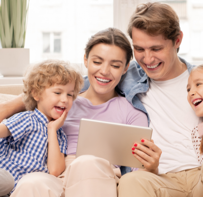 Young joyful casual family of a daughter, father, mother and son sitting on sofa and watching funny video or cartoons on a tablet