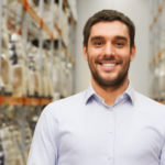 wholesale, logistic, business, export and people concept - happy brunette man in a white shirt with tablet pc computer in a warehouse surrounded by large shelves.