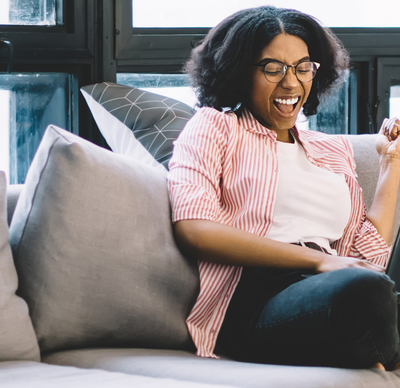 Accomplished woman celebrating something received on her laptop whilst she sits on her grey fabric corner sofa in what looks to be a lounge with a large window wall behind her. The window trim is black. 