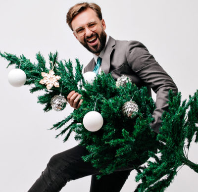 Positive short-haired man in glasses and a suit smiling holding a decorated Christmas tree in front of a white background.