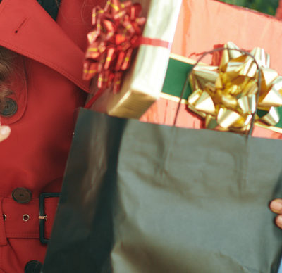 trendy woman in red trench coat from the neck down, with a black shopping bag full of Christmas present boxes sending text message from phone with a Christmas tree behind her