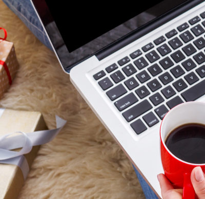 Christmas online shopping above view. The screen is black. Female buyer makes order on laptop, typing with her right hand and holding a hot drink in a red mug, balanced on the edge of the laptop, in her left hand. Cosy blankets in Christmas present bags and brown parcel presents with red and silver ribbons surround her. She is wearing multi coloured fluffy socks, jeans and woolly jumper.
