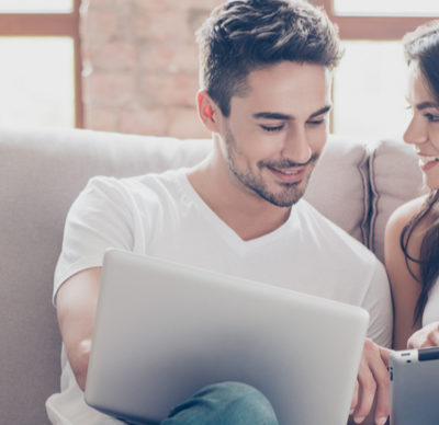 Cheerful happy couple are doing online shopping  on a laptop at home indoors. They are on cosy beige couch in casual clothes, relaxing and buying goods easily
