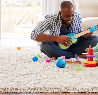 Black father and toddler son playing on a rug in a living room with colourful toys and a play guitar, the father is smiling