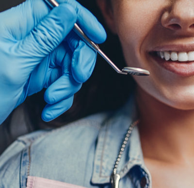 Brunette woman in a dentist clinic, smiling with excellent teeth and male dentist hands either side of her mouth with a dental mirror in the left hand and dental probe in the right hand 