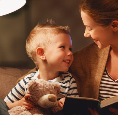 Mother reading her son a book in a dimly lit lounge - evening bedtime story. They are both smiling and the little boy is holding a teddy bear