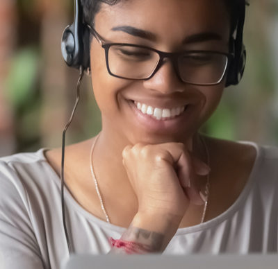 Young woman wearing headphones looking at pc screen, online learning, eLearning. Image is taken from behind laptop. She is smiling. 