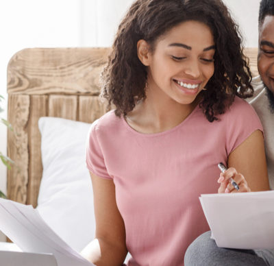 Black man and woman checking on family monthly expenses together, sitting on bed, using laptop and holding papers.